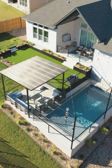 A modern backyard with a small rectangular pool, covered lounge area, artificial turf, and outdoor seating next to a light-colored house.