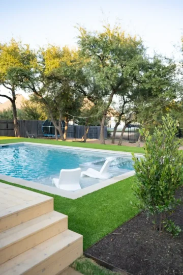 A backyard swimming pool with two white lounge chairs in the shallow end, surrounded by artificial grass, trees, and a wooden deck.