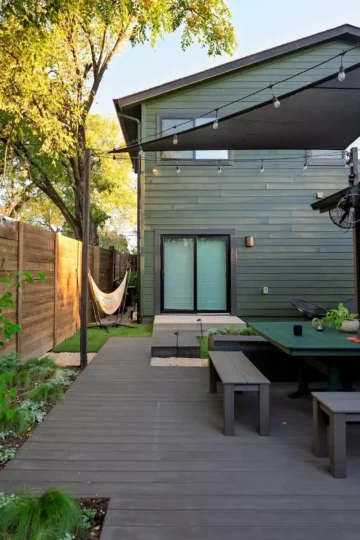 A modern Prospect Hill backyard with a wooden deck, outdoor dining table, bench seating, hammock, string lights, and a two-story house with green siding.
