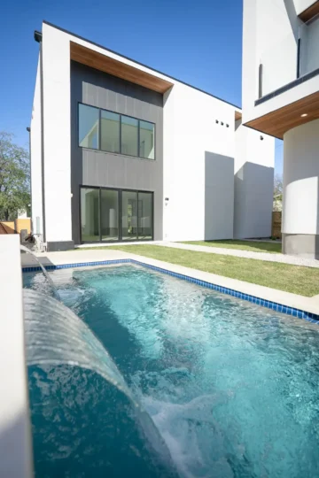 Modern two-story house with large windows and a small rectangular pool in the backyard, featuring a water spout, under a clear blue sky.