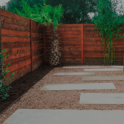 Smooth, rectangular pavers scattered on a bed of gravel surrounded by plants and a wooden fence.