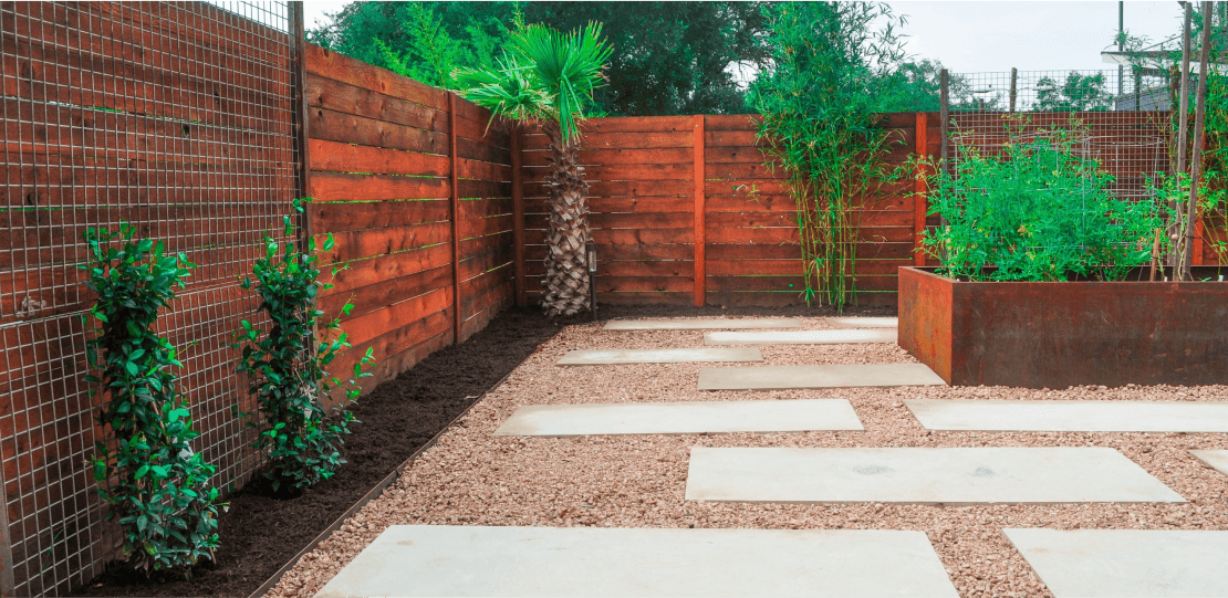 Smooth, rectangular pavers scattered on a bed of gravel surrounded by plants and a wooden fence