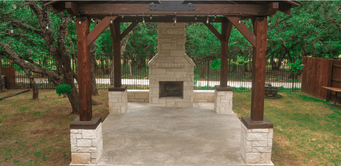 A stone fireplace on a concrete surface covered by a dark wooden pergola.