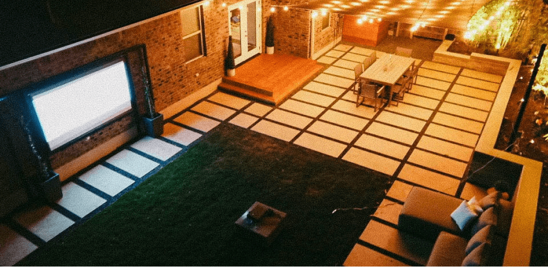 Nighttime birds-eye view of a hardscaping and grass backyard. It is furnished with a blue sectional sofa, projector screen, dining table, and awning with lit up string lights.