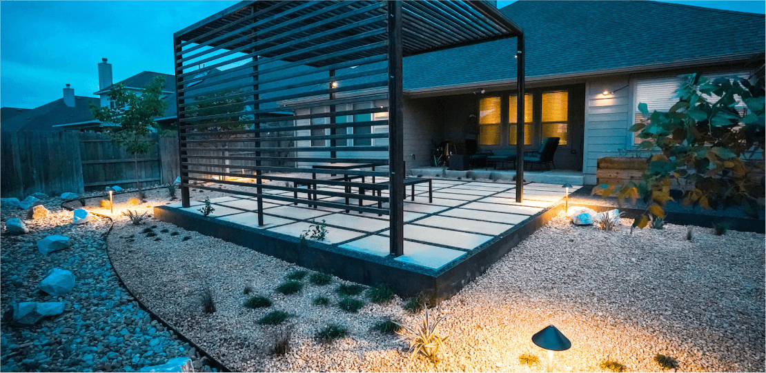 A backyard at dusk with a metal pergola overtop a table and 2 benches on hardscaping tiles.