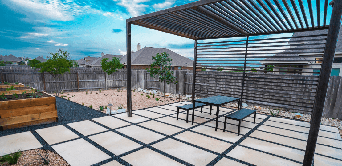 A backyard with a metal pergola overtop a table and 2 benches on hardscaping tiles.