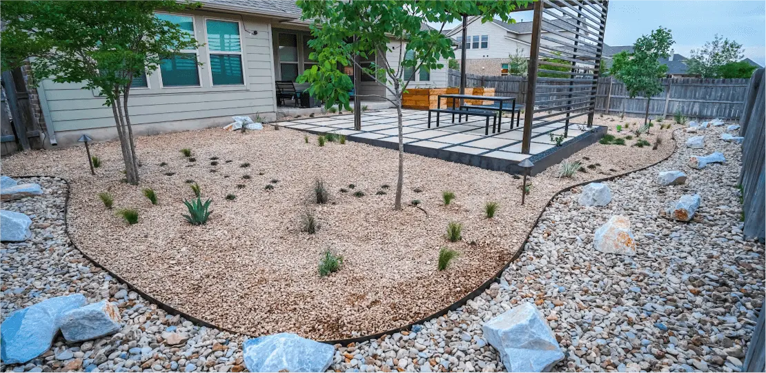 A backyard filled with gravel, small plants, and trees in front of a pergola.