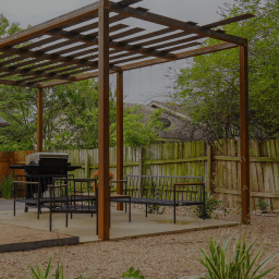 A gravel backyard with a metal pergola covering a grill, table, and chairs.