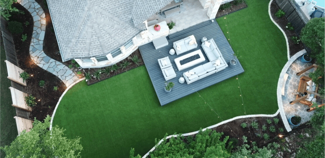A birdseye view of a backyard patio furnished with chairs, sofas, and a fire pit surrounded by grass.