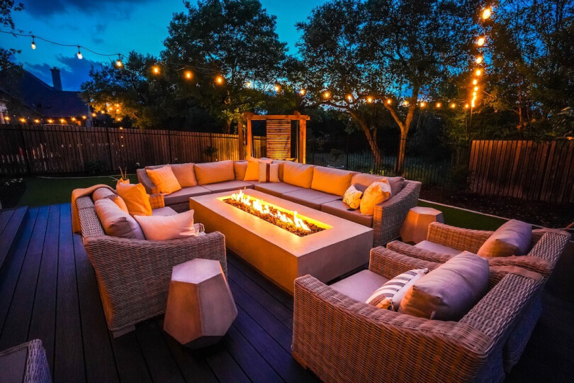 Outdoor sofas and chairs surrounding a rectangular fire pit with string lights hanging above.