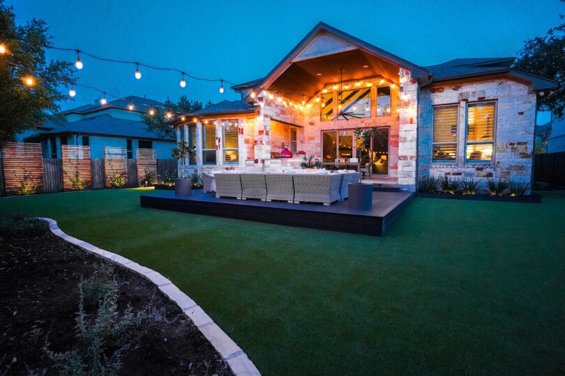 A grassy backyard with a raised living area illuminated by string lights.