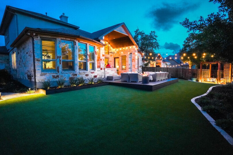 A grassy backyard with a raised living area illuminated by string lights.