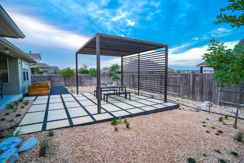 A backyard with a metal pergola on hardscaping tiles surrounded by gravel.