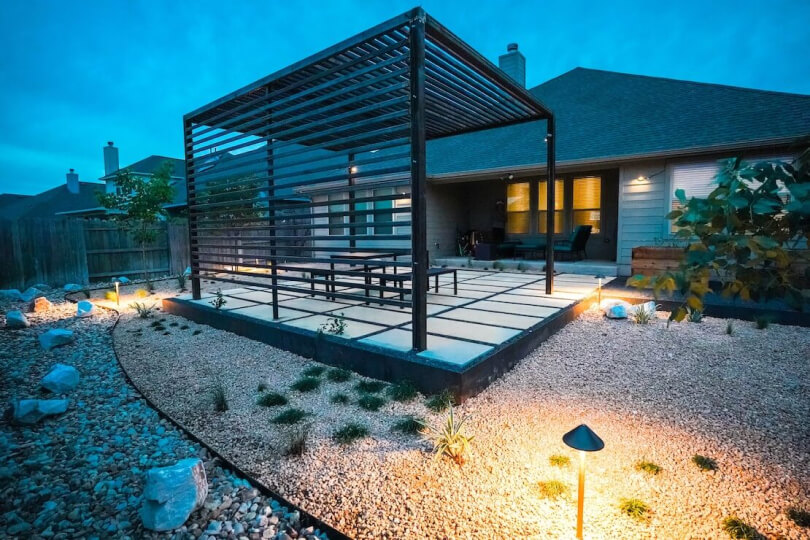Rear view of a backyard with a metal pergola on hardscaping tiles surrounded by gravel.