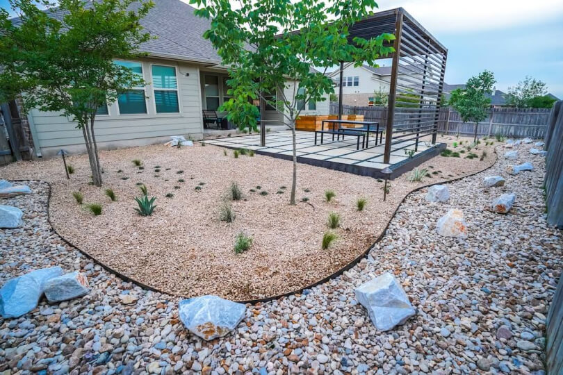 A backyard filled with gravel, small plants, and trees in front of a pergola.