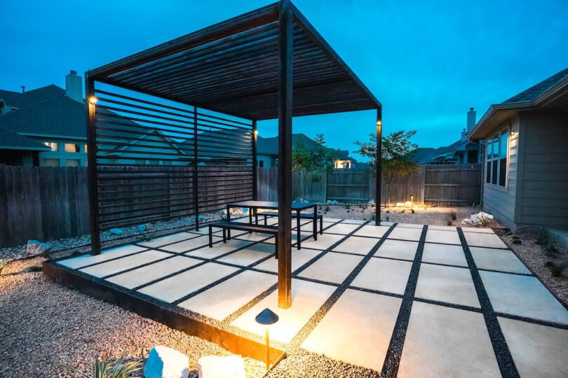 A backyard at dusk with a metal pergola overtop a table and 2 benches on hardscaping tiles.