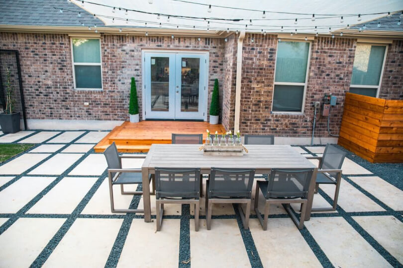 Backyard hardscaping of rectangular tiles with gravel in between shown with a table and 8 chairs atop it.