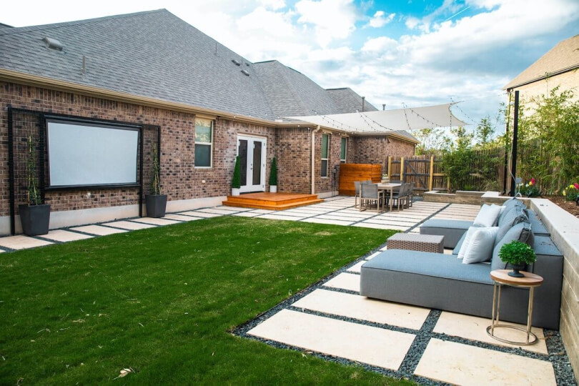 The backyard of a house with grass and hardscaping furnished with a blue sectional sofa and a projector screen.