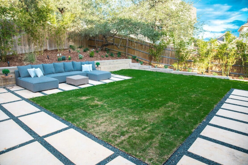 Grass in a backyard surrounded by hardscaping tiles and furnished with a blue sectional sofa.