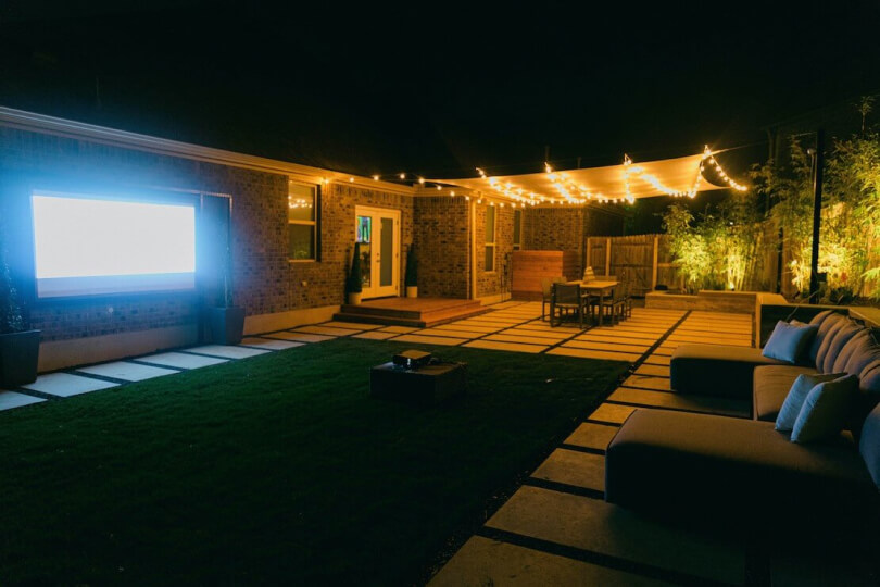 Nighttime view of a hardscaping and grass backyard. It is furnished with a blue sectional sofa, projector screen, dining table, and awning with lit up string lights.