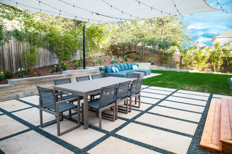Backyard with a mix of hardscaping tiles and grass. A table with 8 chairs sits on the hardscaping covered with a white awning.