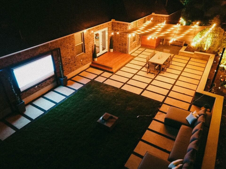 Nighttime birds-eye view of a hardscaping and grass backyard. It is furnished with a blue sectional sofa, projector screen, dining table, and awning with lit up string lights.