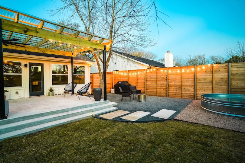 A backyard with a concrete patio covered by a wooden pergola, gravel, and a small pool.