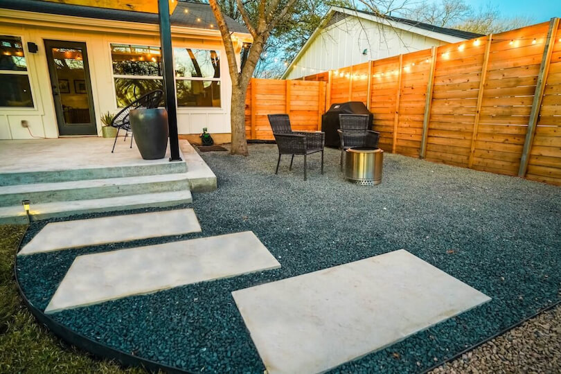 3 paved tiles on gravel leading to a patio area. On the right there are 2 chairs around a fire pit.