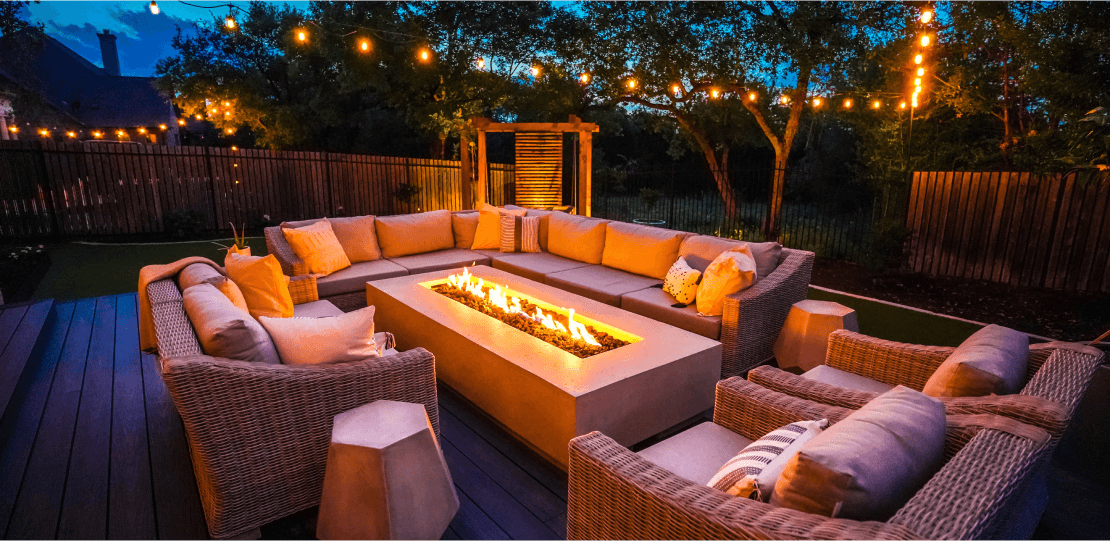 Outdoor sofas and chairs surrounding a rectangular fire pit with string lights hanging above.