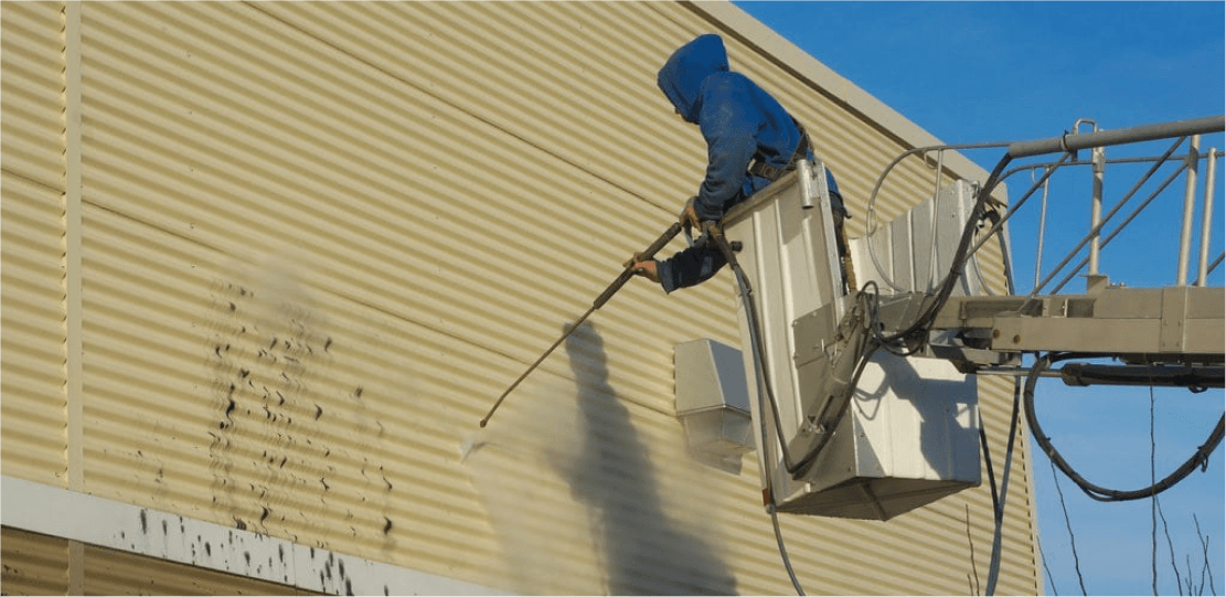 A person wearing a blue hoodie in a lift power washing the side of a house.