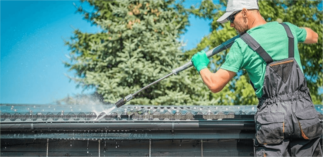 A person in a green shirt and overalls power washing a roof.