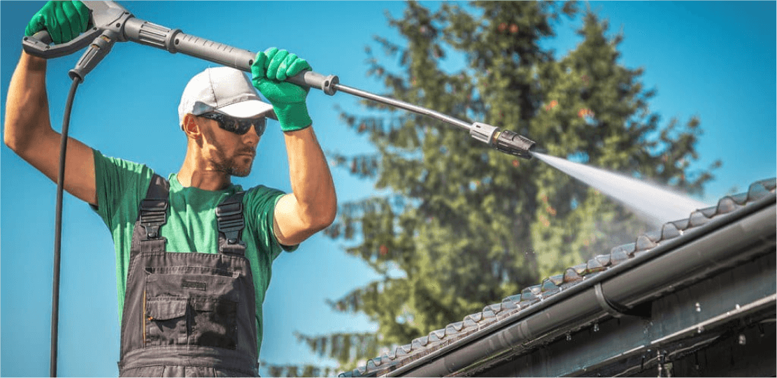 A person in a green shirt and overalls power washing a roof.