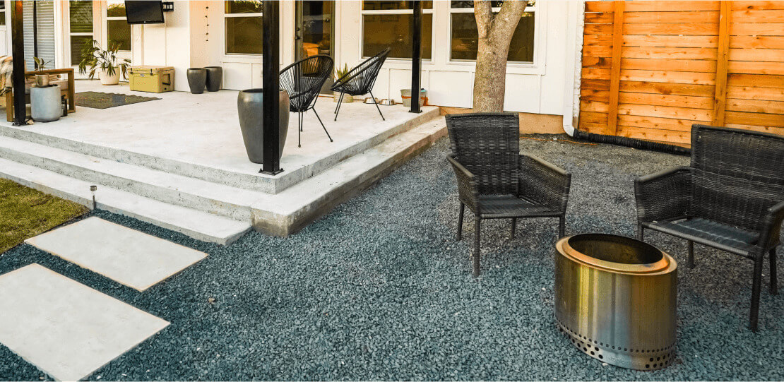 3 paved tiles on gravel leading to a patio area. On the right there are 2 chairs around a fire pit.