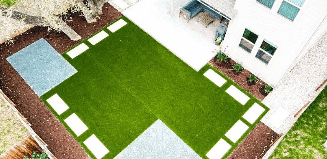 Birdseye view of a mostly empty grassy backyard. Rectangular paved stones line the edges of the grass.