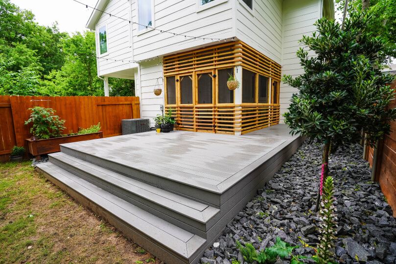 Grey deck with a couple steps leading up to it. Atop the porch, there is a screened in porch with light brown wooden archways around it.