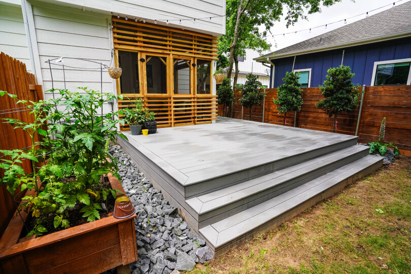 Grey deck with a couple steps leading up to it; on the left side, there are grey rocks and a cedar planter. Atop the porch, there is a screened in porch with light brown wooden archways around it.