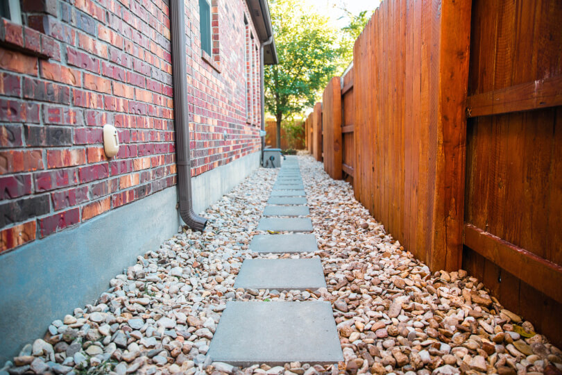 Square cement pavers laid in a path between the side of a house and a fence. There are large pebbles between the pavers.