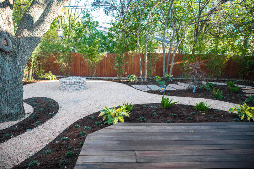 Backyard with a wooden deck, trees, gravel path, and areas of mulch with newly planted shrubs.