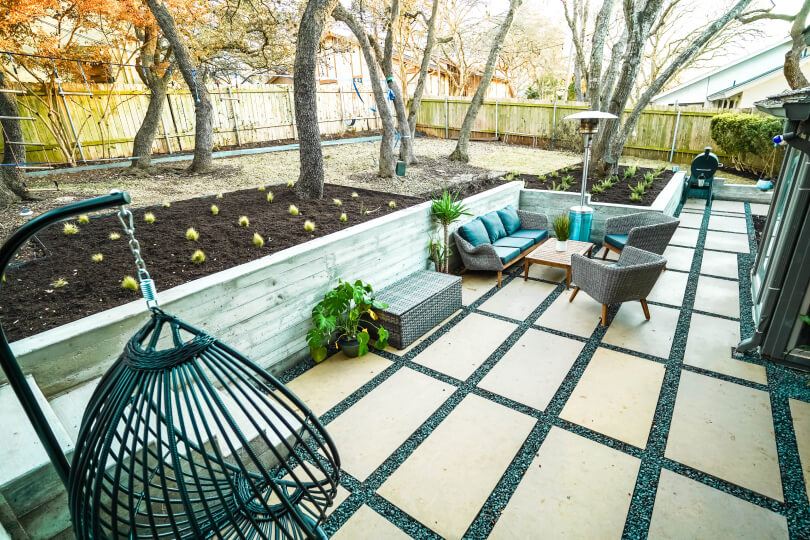 Backyard with a sunken area tiled with light-colored rectangular pavers with grey gravel between them. Inside the sunken area, there is a seating area with a wicker sofa and chairs and a black swing. Up the steps from the sunken area is a concrete planter with evenly spaced sprouts and a grassy area with trees behind it.