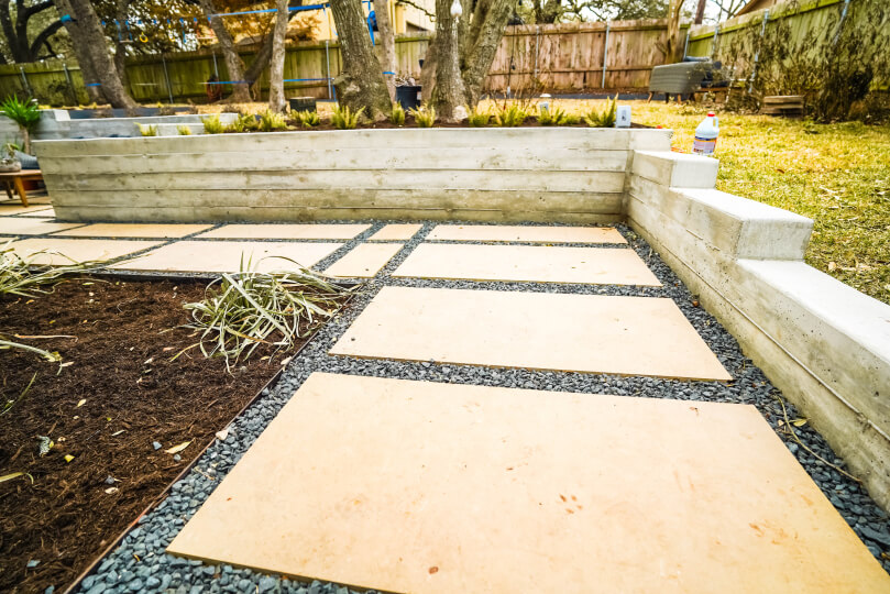 Close up of a sunken area of a backyard tiled with lightly-colored rectangular pavers lined with grey gravel. Along the edges of the sunken area is a low concrete wall and a grassy area with trees on the other side of the wall.
