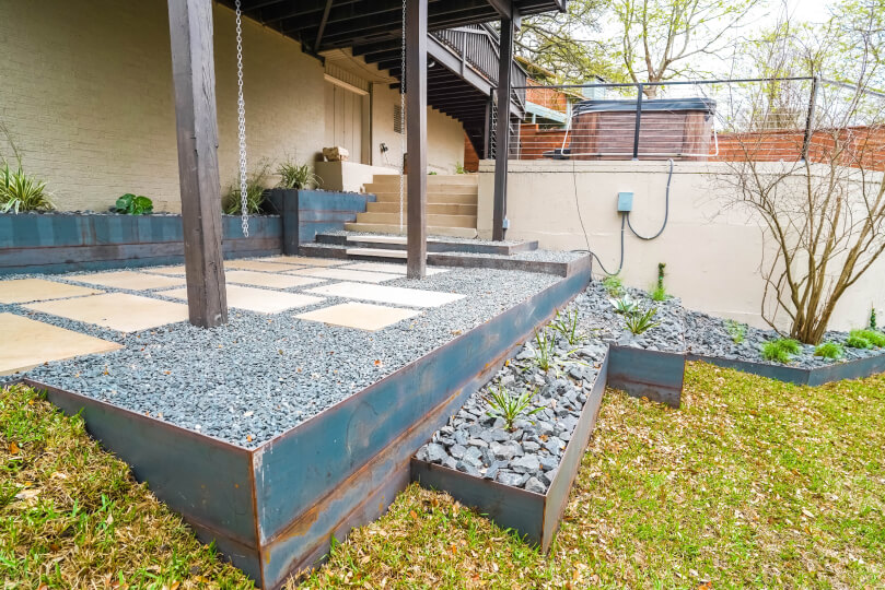 Tiered flower beds filled with grey pebbles leading up to a landing with lightly-colored rectangular pavers lined with smaller grey pebbles. From the landing, stairs lead up to a deck area.