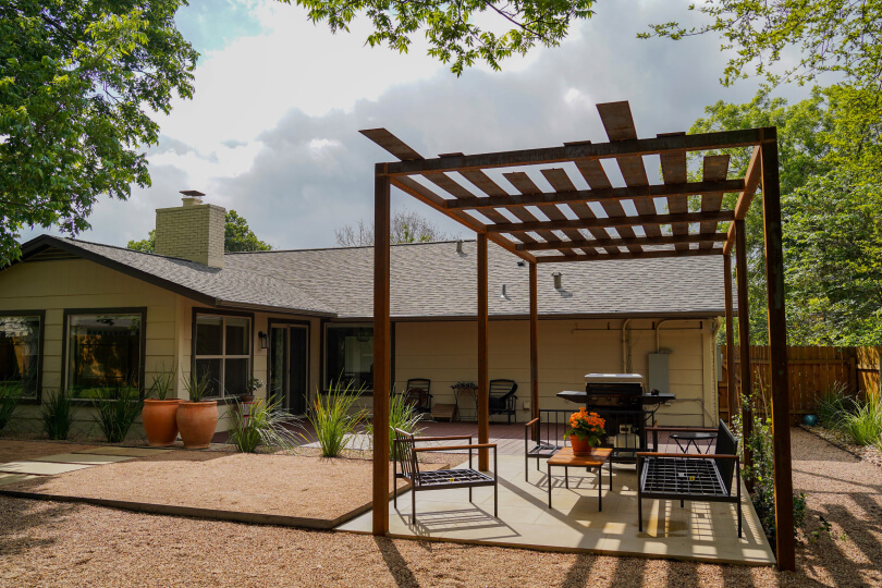 Metal pergola with a grill and chairs underneath. In the background, a house is visible.