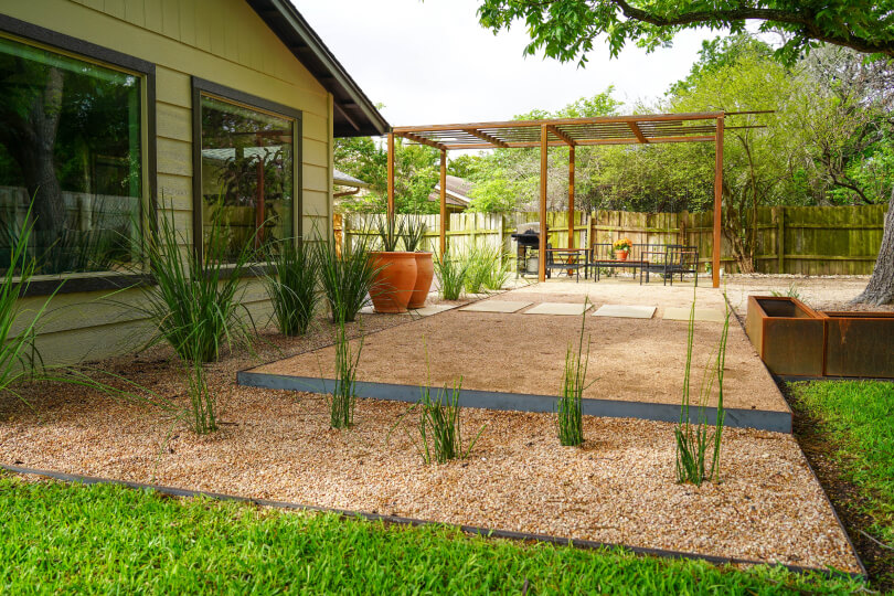 Backyard with a mix of gravel and grass with sparse plants in the gravel. In the background, a metal pergola is visible.