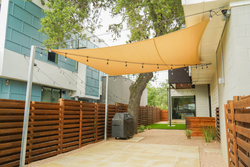 Yard with a patch of lightly-colored stone with a grill on it and a shade sail with string lights above it.