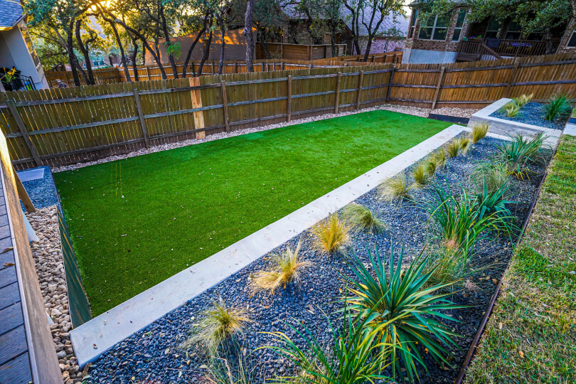 Backyard with lush green grass, cement planters with spaced out plants and grey gravel between them, and a light wooden fence with vertical slats along the edge of the yard.