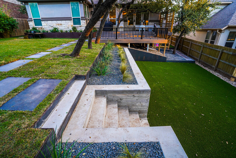 Lush green grass leading up to raised cement planters with stairs cut into the middle of the planters. The stairs lead to a raised grassy area with trees.