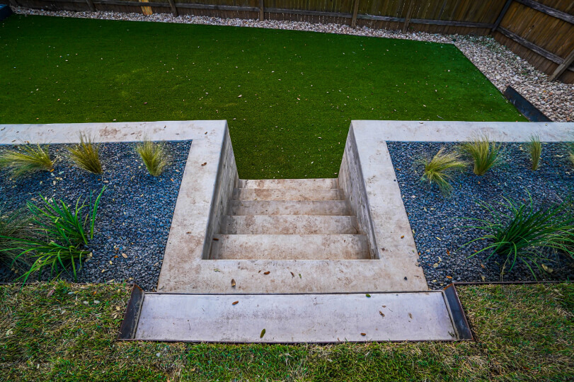 Aerial view of cement planters with grey gravel and small spiky plants. There are cement stairs cut into the middle of the planters. The stairs lead to a lush grassy area at their base.