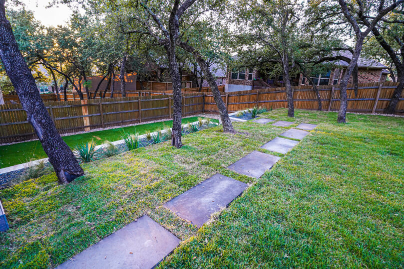 Grassy area of a backyard with light rectangular pavers laid out in a path next to a few spaced out trees.