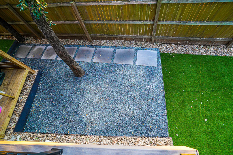 Aerial view of a patch of grey gravel with square pavers laid in a path next to a light wooden fence. The path leads to a patch of lush green grass.
