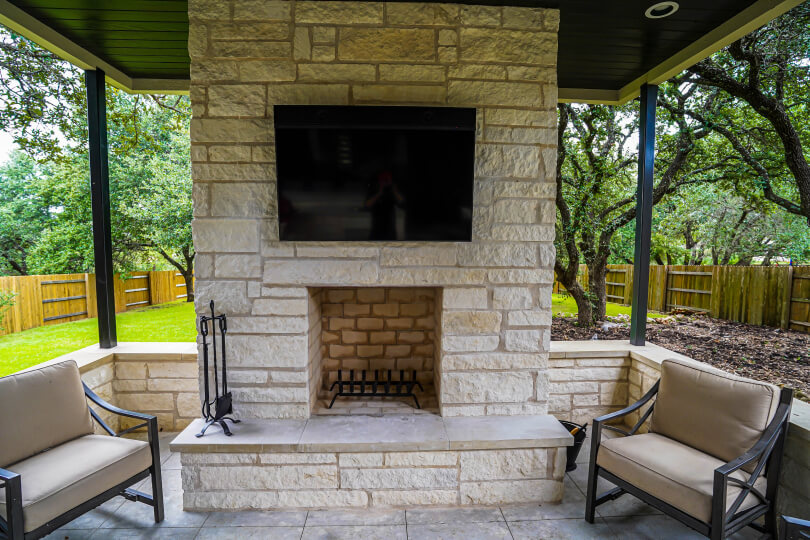 Light stone fireplace with a TV mounted above it and 2 chairs next to the fireplace.